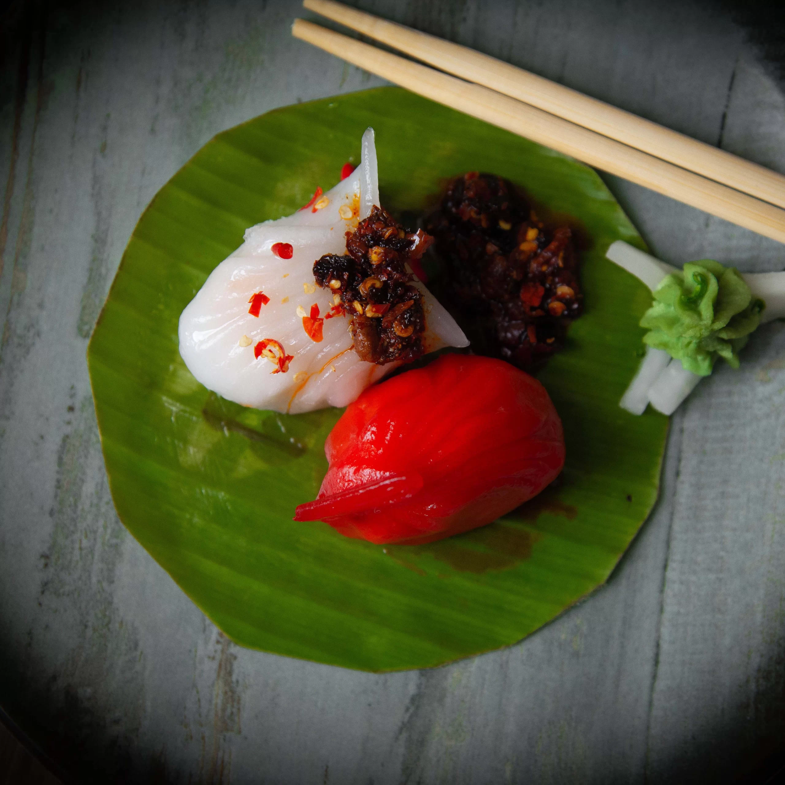 Photography Portfolio Contemporary Asian dim sum dumpling with chili oil and red ginger flower on banana leaf plate with chopsticks - Content Crest Media modern Asian cuisine photography portfolio Ahmedabad Gujarat